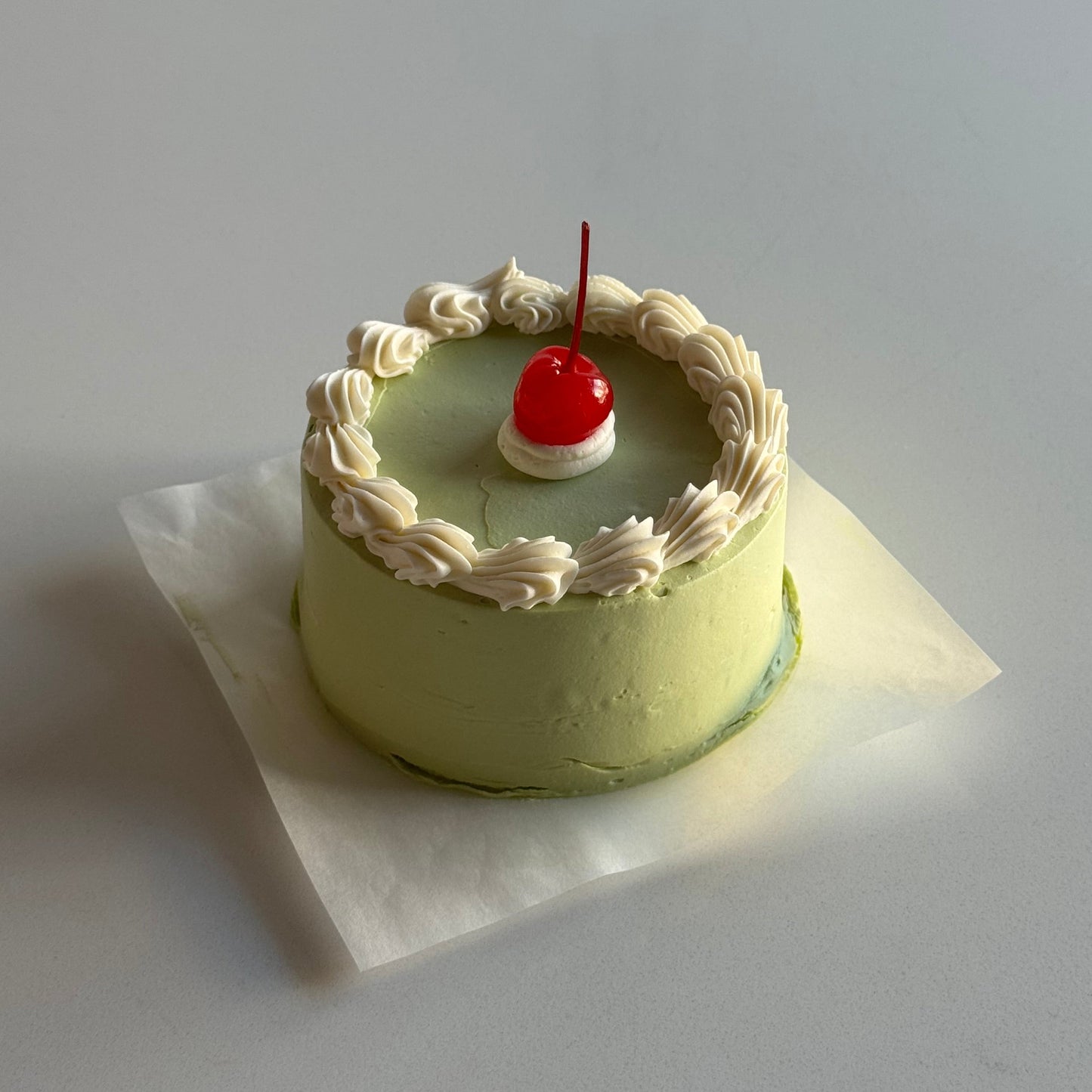 Small cake with white frosting and a cherry on top, placed on a white surface.