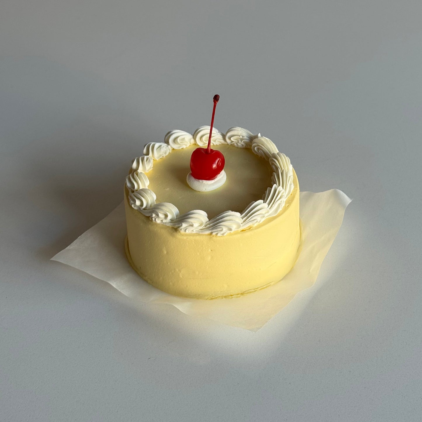Small round cake with white frosting and a red cherry on top, placed on a white paper against a light gray background.
