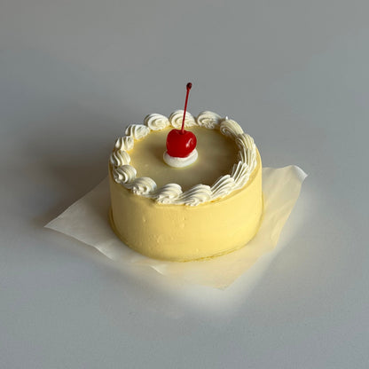 Small round cake with white frosting and a red cherry on top, placed on a white paper against a light gray background.
