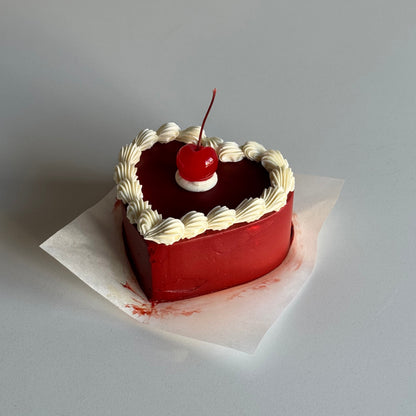 Small red cake with white frosting and a cherry on top, placed inside a triangular cutout of a larger cake.