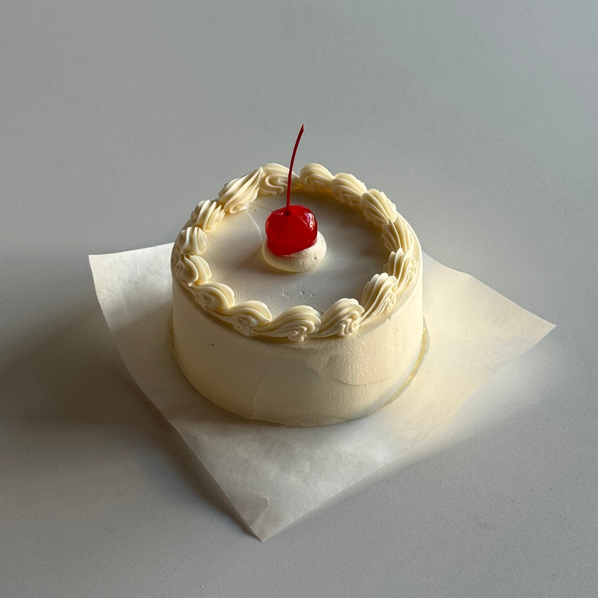 Small round cake with white frosting and a red cherry on top, placed on a white surface.