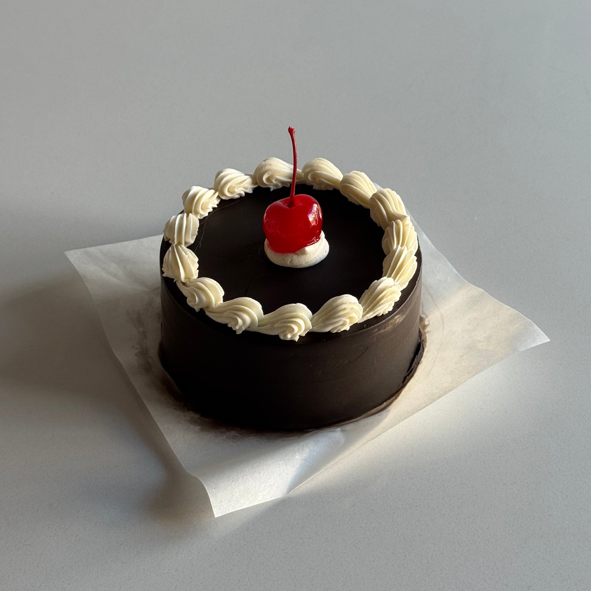 Small chocolate cake with white frosting and a red cherry on top, placed on a white surface.